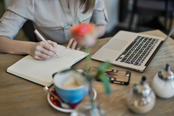 Femme assise à un bureau avec un ordinateur, des lunettes et une tasse de café qui sont posés sur la table et qui écrit dans un cahier.