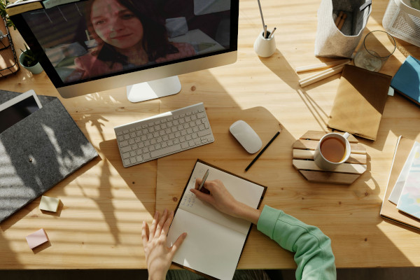 Personne assise à son bureau et qui prend des notes dans un cahier lors d'une visio avec une femme.